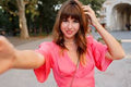 Young woman with brown hair and bangs taking a selfie outdoors wearing a bright pink top.